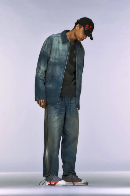 Full-length studio shot of a person wearing an ombré denim jacket, jeans, t-shirt, sneakers, and baseball cap against a light backdrop.