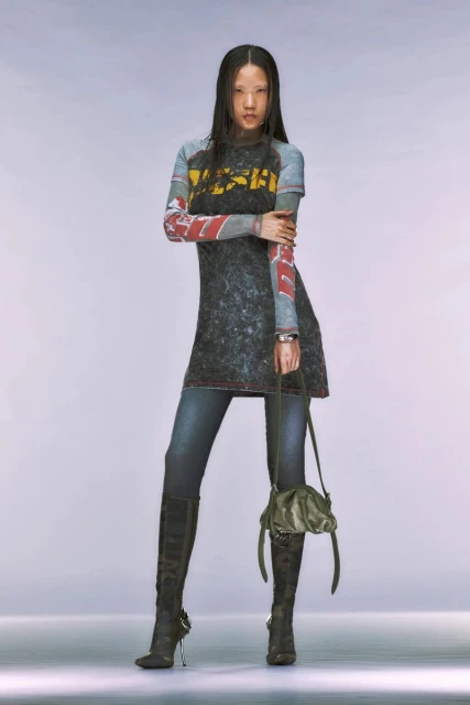 Asian female model stands in a studio wearing a printed t-shirt dress, skinny jeans, heeled boots, and carrying a slouchy bag.