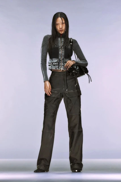 A full shot captures a stylish Asian woman in a gray top, black vest, and cargo pants, accessorized with a black bag, set against a bright studio backdrop.