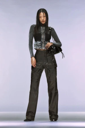 A full shot captures a stylish Asian woman in a gray top, black vest, and cargo pants, accessorized with a black bag, set against a bright studio backdrop.