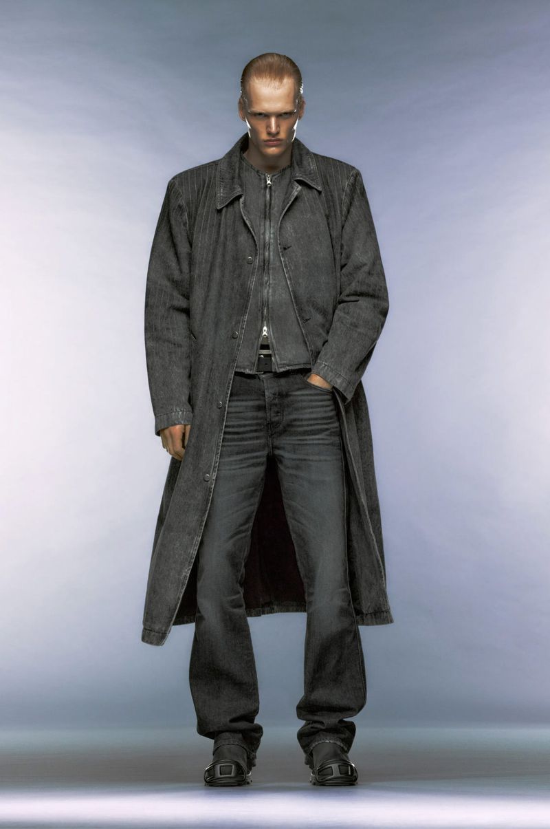 Full-length studio shot of a man in a long denim coat, matching jeans and black boots against a cool grey gradient background.