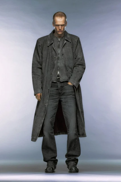 Full-length studio shot of a man in a long denim coat, matching jeans and black boots against a cool grey gradient background.