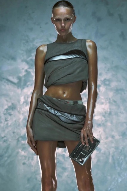 A fashion photo featuring a model wearing a gray sleeveless top and skirt with metallic accents, holding a silver clutch against a textured backdrop.