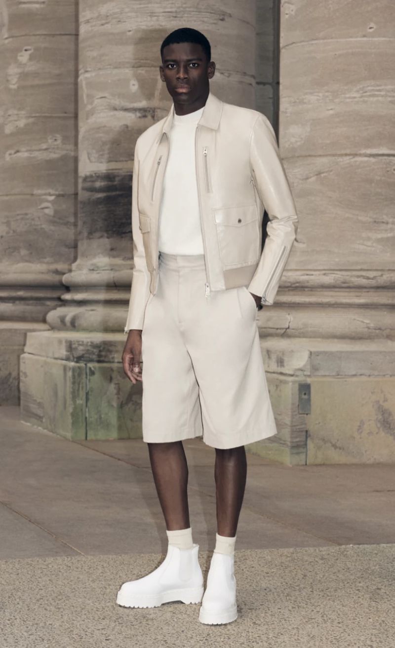An African American man models light beige outfit in an architectural setting, featuring a jacket, shorts, a white t-shirt, and white boots.