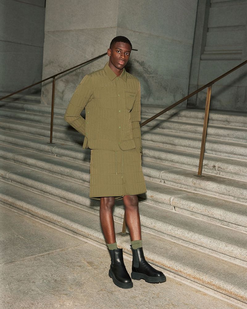 A young Black man models olive-green jacket and skirt with black boots on stone steps, creating a fashion editorial shot.