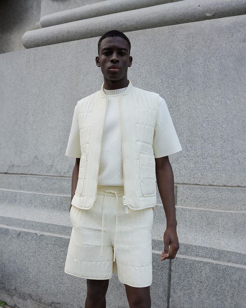 Portrait of a young man in a matching quilted vest and shorts ensemble, posing outdoors against a textured concrete background.