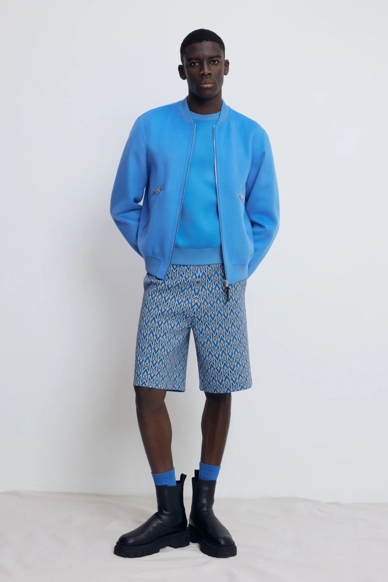 A tall, dark-skinned man in a blue jacket, blue shirt, and patterned shorts stands in front of a white background in a studio shot.