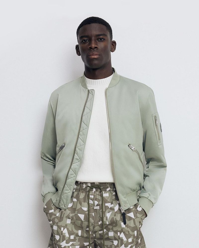 Studio shot of a Black male model in a light green bomber jacket and patterned pants against a plain background, showcasing fashion and style.