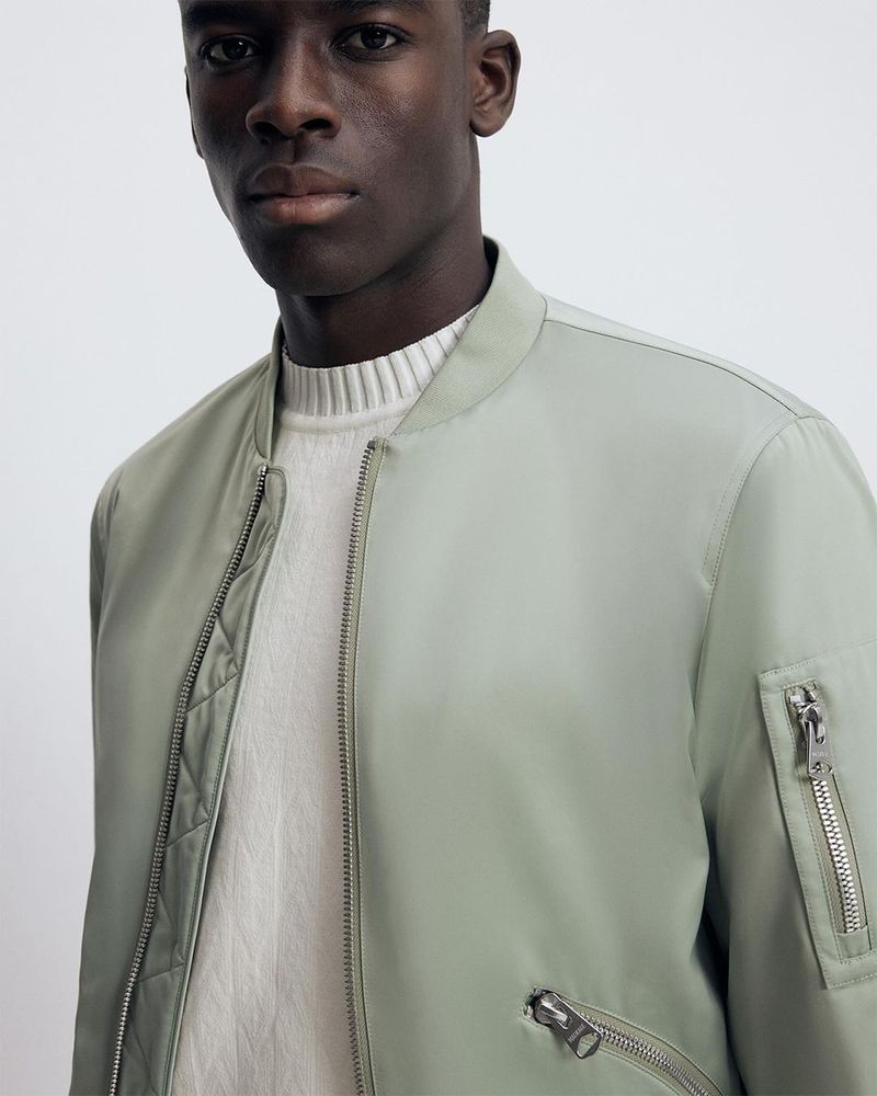 Close-up studio shot of a dark-skinned adult male wearing a pale green bomber jacket and a cream-colored knit sweater. Fashion advertisement.