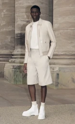 Black man wearing cream leather jacket and matching shorts standing before classical stone pillars