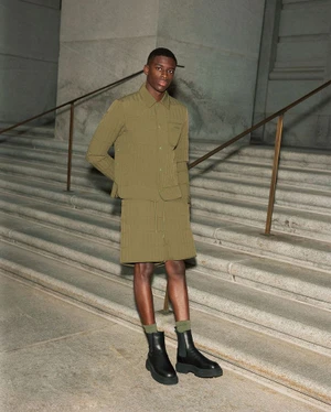 Black man standing on stone stairs wearing an olive quilted shirt, matching shorts, and black boots.