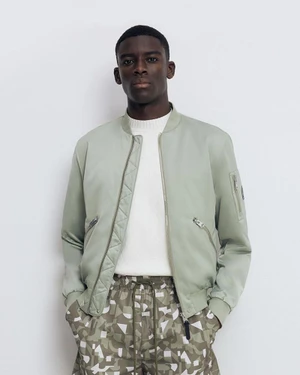 Black man wearing a sage green bomber jacket over a white sweater against a plain white studio background.