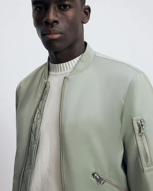 Black man wearing a sage green bomber jacket and ribbed white sweater against a plain white background