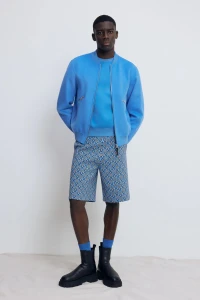 Model wearing a blue jacket and patterned shorts with black boots against a white studio background