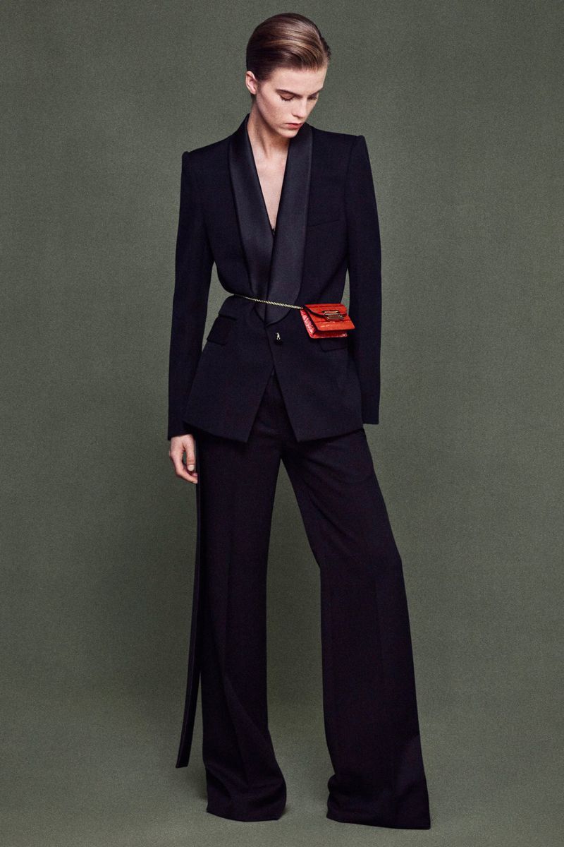 Fashionable woman in black suit with red waist bag posing against a muted green background in a studio setting, full body shot.