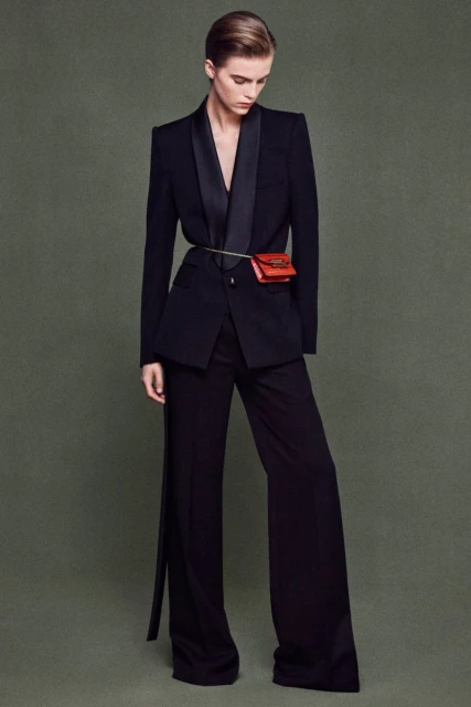 Fashionable woman in black suit with red waist bag posing against a muted green background in a studio setting, full body shot.