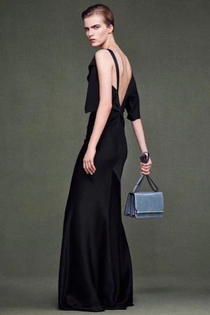 An adult female model with short hair poses in a black, floor-length gown and holds a blue purse against an olive green background.