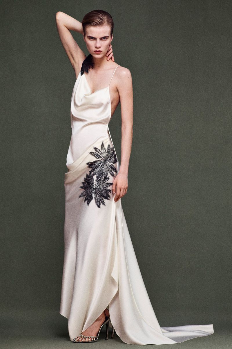 Woman poses in white gown with flower detail in a studio. The model's arm is raised, highlighting the open-back dress design.