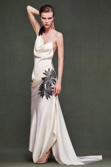 Woman poses in white gown with flower detail in a studio. The model's arm is raised, highlighting the open-back dress design.