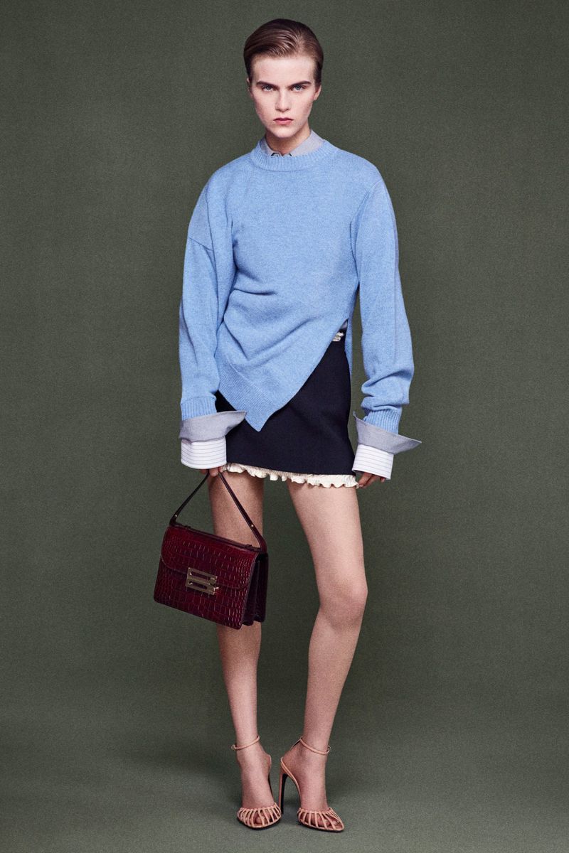 Full shot of a female model in a light blue sweater, black skirt, and high heels, carrying a burgundy purse against a green background.