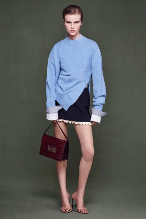 Full shot of a female model in a light blue sweater, black skirt, and high heels, carrying a burgundy purse against a green background.