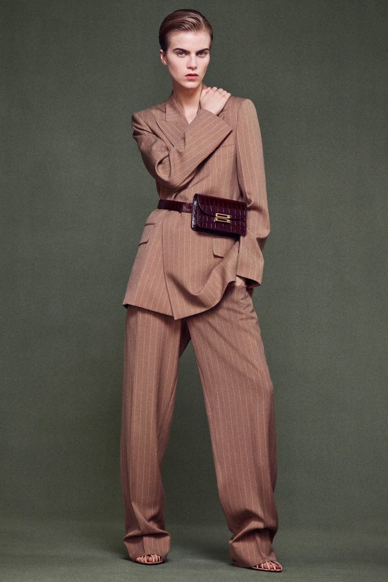 Fashion shot of a woman in a tan pinstripe suit with wide-leg pants, paired with a brown belt bag, set against a neutral background.