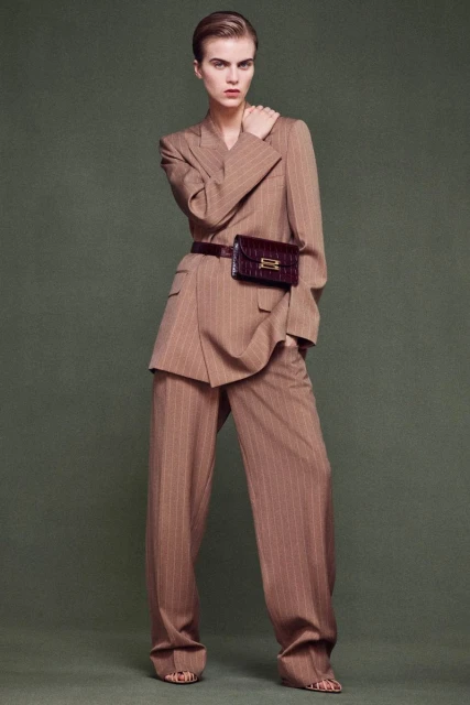 Fashion shot of a woman in a tan pinstripe suit with wide-leg pants, paired with a brown belt bag, set against a neutral background.