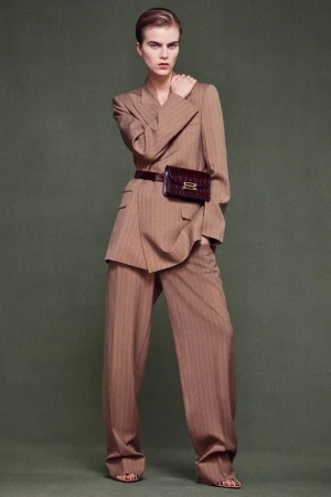 Fashion shot of a woman in a tan pinstripe suit with wide-leg pants, paired with a brown belt bag, set against a neutral background.
