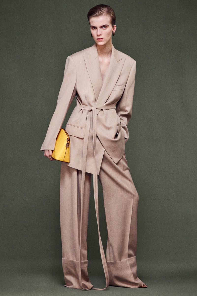 Fashion editorial shot: A tall woman in a beige suit and yellow bag poses against a green background in a full-length shot.