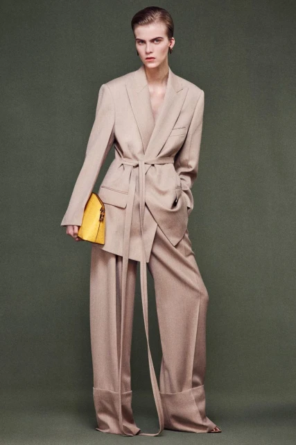 Fashion editorial shot: A tall woman in a beige suit and yellow bag poses against a green background in a full-length shot.