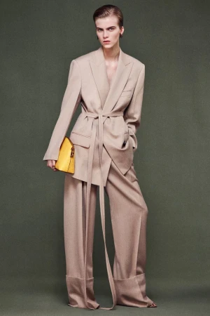 Fashion editorial shot: A tall woman in a beige suit and yellow bag poses against a green background in a full-length shot.