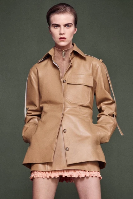 Fashionable young woman in a tan leather jacket and matching skirt poses against a muted green background in a studio setting.
