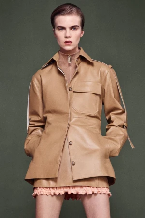 Fashionable young woman in a tan leather jacket and matching skirt poses against a muted green background in a studio setting.