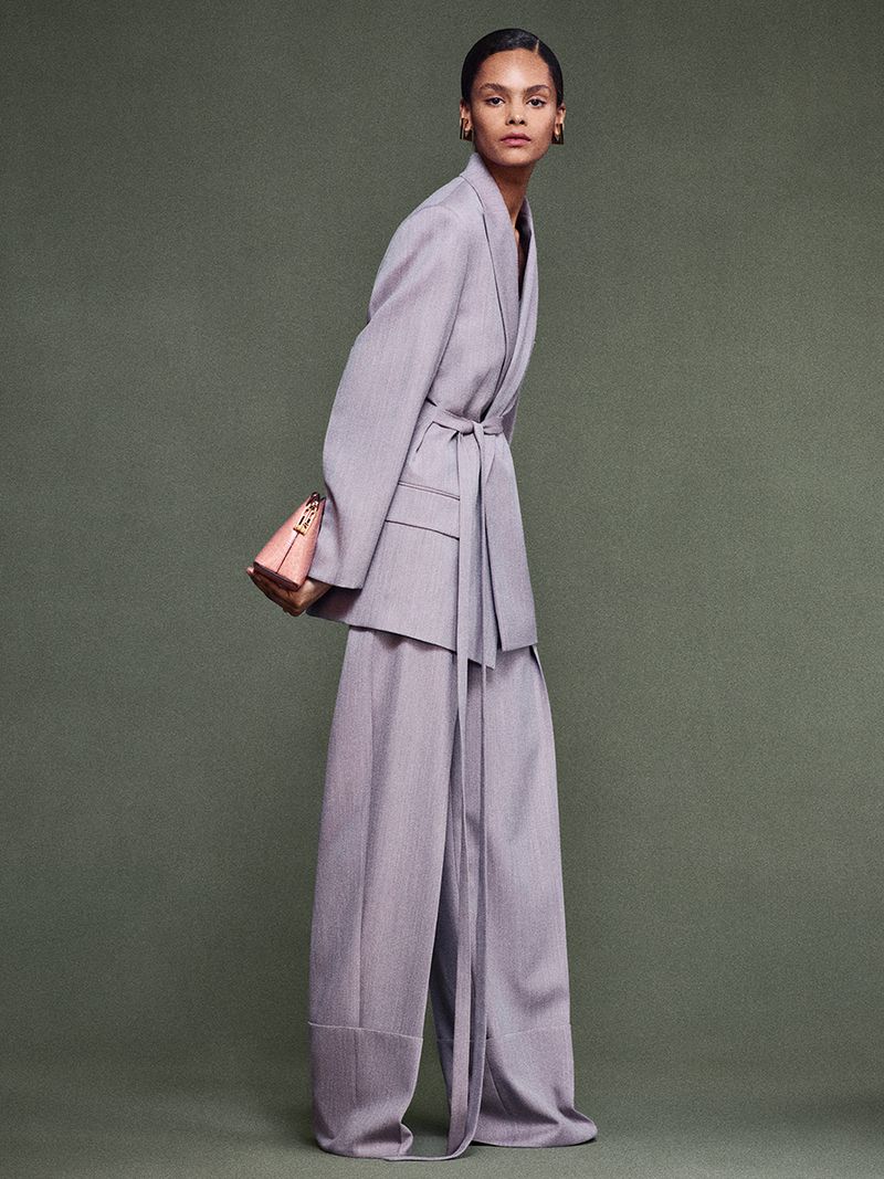 Fashion model in a light purple blazer with matching trousers, accessorized with a structured purse, posing in a studio setting.