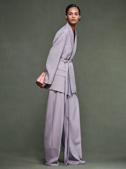 Fashion model in a light purple blazer with matching trousers, accessorized with a structured purse, posing in a studio setting.