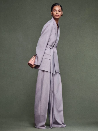 Fashion model in a light purple blazer with matching trousers, accessorized with a structured purse, posing in a studio setting.