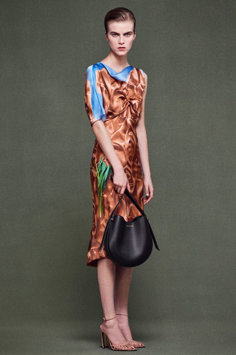 Elegant woman in a patterned midi dress holding a black bucket bag, posing against a solid green background, showcasing fashion and style.