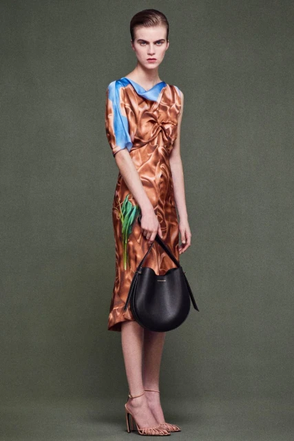 Elegant woman in a patterned midi dress holding a black bucket bag, posing against a solid green background, showcasing fashion and style.
