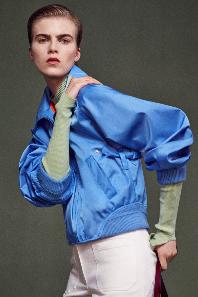 Fashion editorial shot of a young adult woman posing in a light blue jacket and white pants against a neutral studio backdrop.