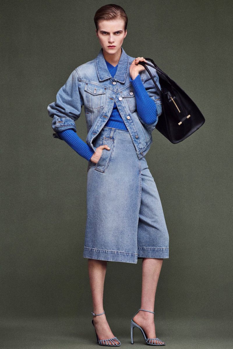 Fashion model wearing blue denim jacket and culottes, holding a black leather tote bag, posing against a neutral olive green background.