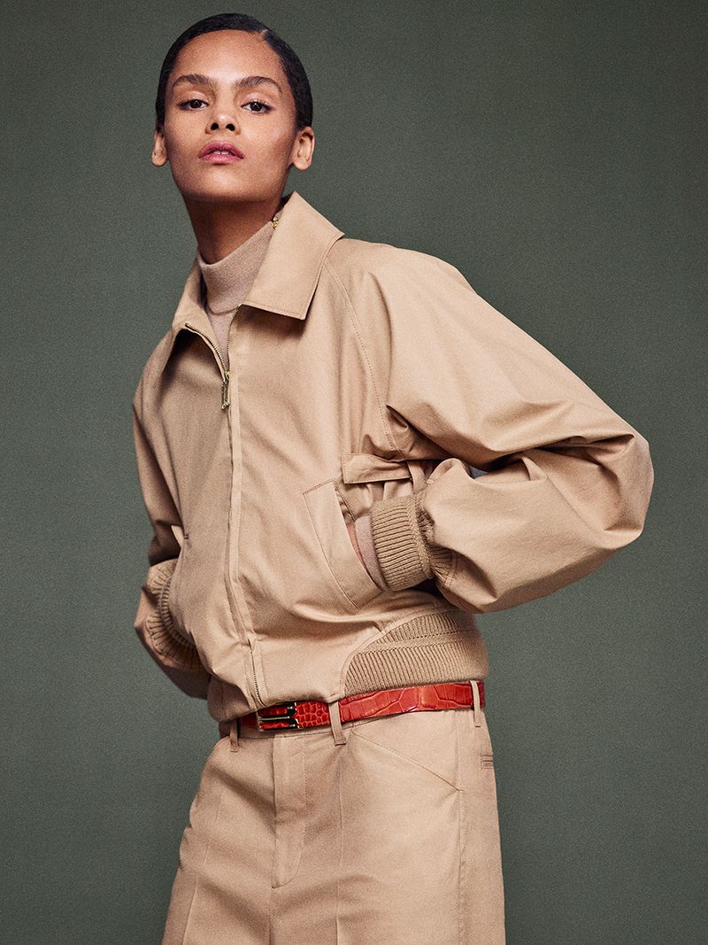Fashionable woman in a studio shot wearing a tan jacket, pants, and turtleneck with a red belt, exuding a modern editorial vibe.