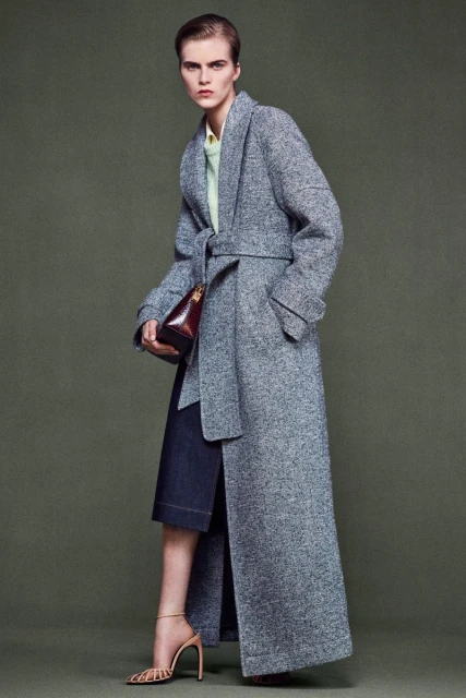 A woman models a full-length grey wool coat cinched with a belt over a dark denim skirt, accessorized with a brown purse and strappy heels.