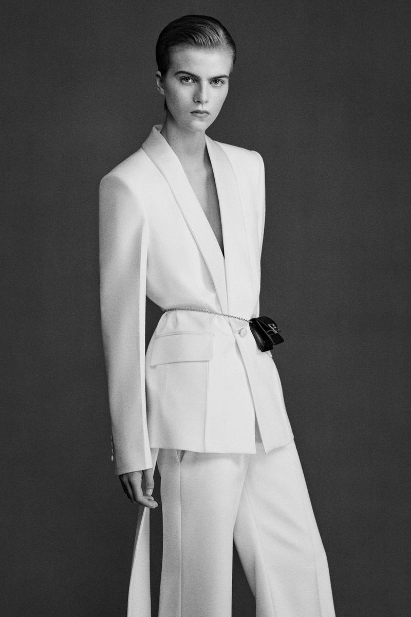 Monochrome fashion shot of a female model with short hair wearing a white pantsuit with a black purse on a belt against a grey backdrop.