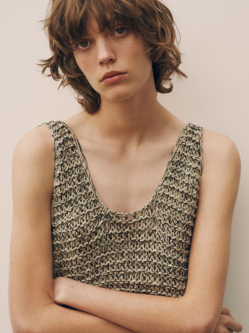 Close-up of a female model with short wavy hair wearing a knitted tank top against a neutral background in a studio setting.