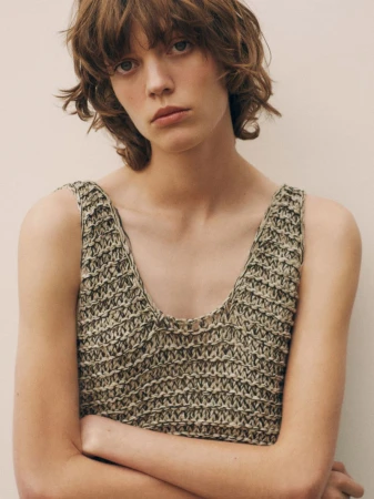 Close-up of a female model with short wavy hair wearing a knitted tank top against a neutral background in a studio setting.