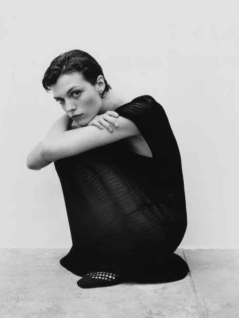 Monochrome portrait of a female model in a black dress, squatting indoors against a white background in a fashion editorial style.