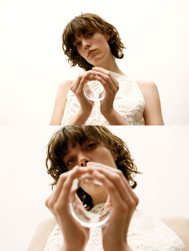 Young woman in a white tank dress holds a glass ring up to the camera in a high-angle studio shot with a bright neutral background.