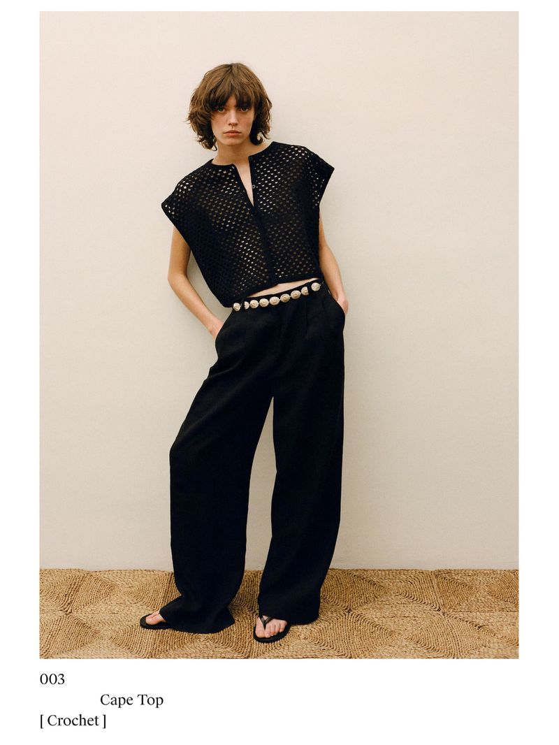 A woman poses in a black crochet top, black pants, and sandals against a neutral background, exuding a relaxed yet stylish fashion aesthetic.