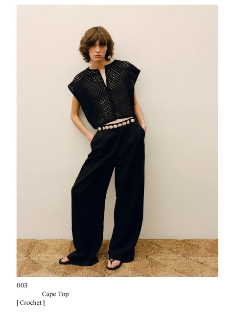 A woman poses in a black crochet top, black pants, and sandals against a neutral background, exuding a relaxed yet stylish fashion aesthetic.
