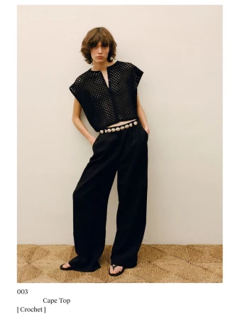 A woman poses in a black crochet top, black pants, and sandals against a neutral background, exuding a relaxed yet stylish fashion aesthetic.